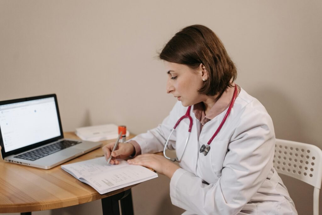 Photographie de profil d'une femme médecin en blouse blanche avec un stéthoscope rose, écrivant à la main dans un carnet sur son bureau, à côté d'un ordinateur portable ouvert. Ce visuel illustre le processus de validation administrative nécessaire après avoir demandé un arret maladie en ligne.